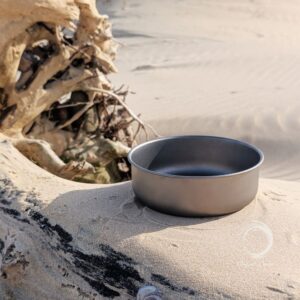 Deep titanium camping bowl set on driftwood in a sandy coastal landscape
