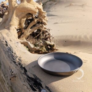 Titanium camping plate resting on driftwood in the sand at a coastal campsite