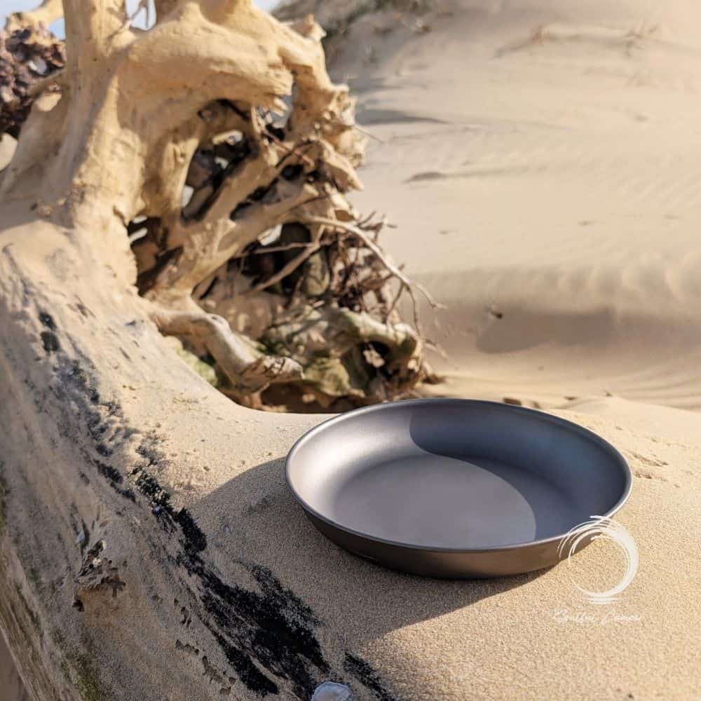 Titanium camping plate resting on driftwood in the sand at a coastal campsite