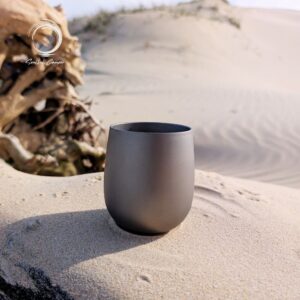 Pure titanium camping cup placed on a sand dune with driftwood, ideal for lightweight outdoor adventures.