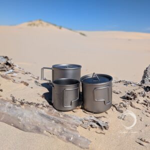 Titanium camping mugs in three sizes from the Ti-3 Mug Stack displayed on driftwood in the Anna Bay sand dunes, showing lightweight nesting outdoor mug set.